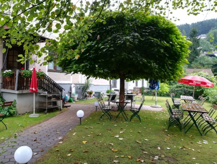 Ein gemütlicher Gastgarten mit Tischen und Stühlen unter einem großen Baum, umgeben von grüner Landschaft.