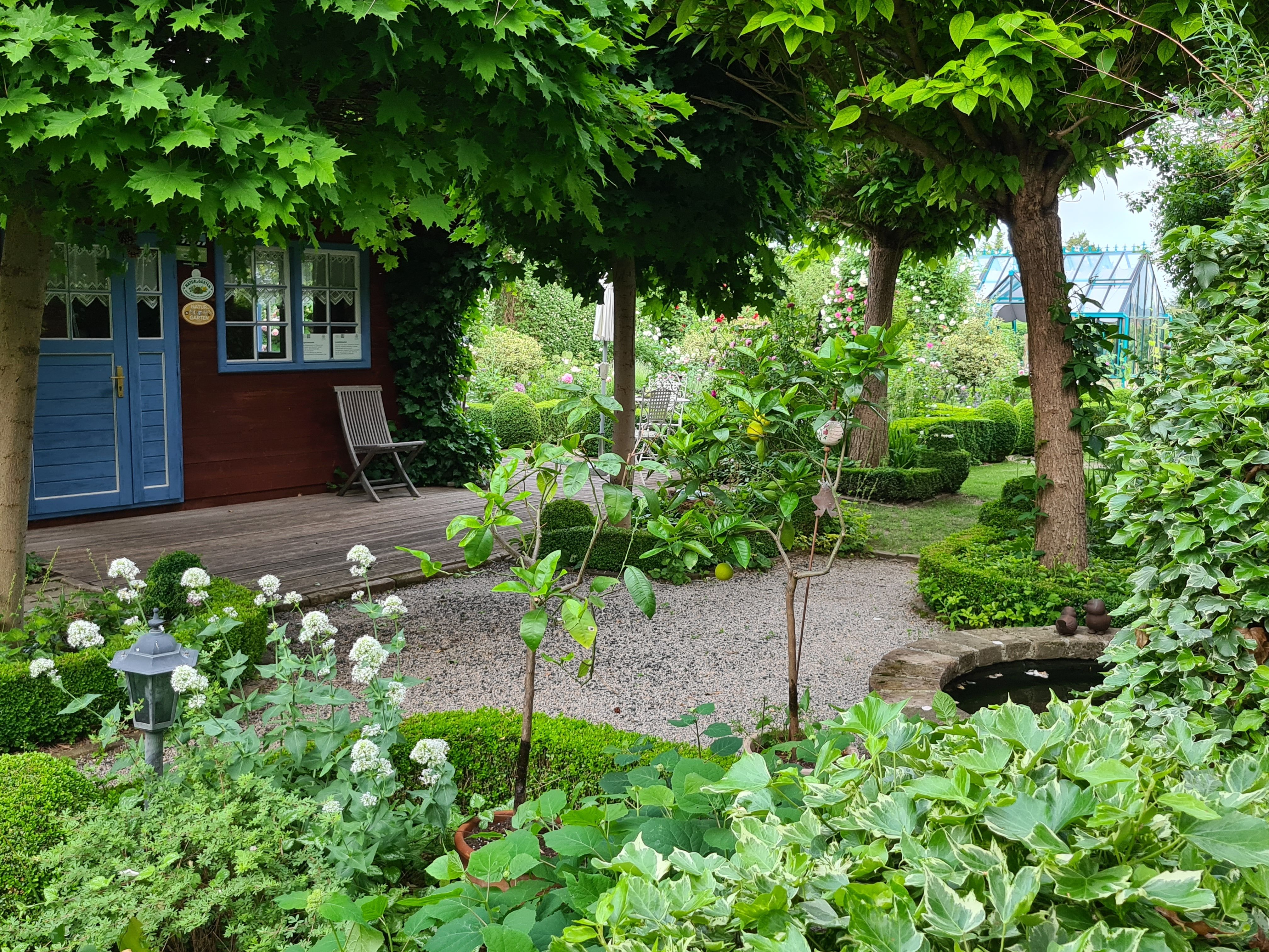 Ein idyllischer Garten mit einem kleinen Holzhaus, umgeben von Bäumen und Pflanzen, mit einem Kiesweg und einem kleinen Teich.