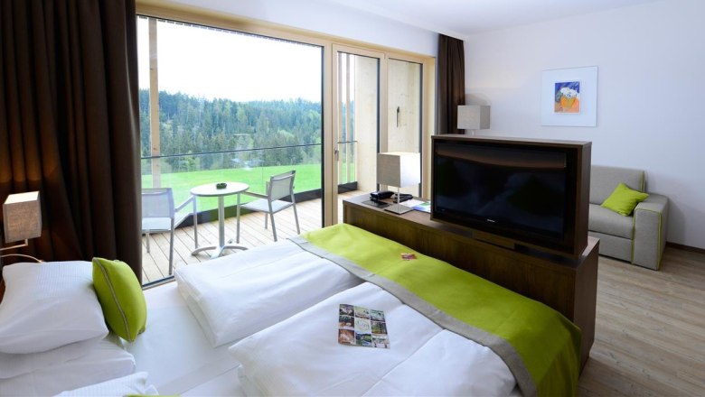 Modernes Hotelzimmer mit Doppelbett, Fernseher, Sofa und Balkon mit Waldblick.