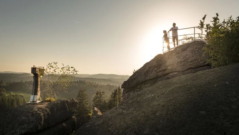 Zwei Personen stehen auf einem Felsen mit Geländer, im Hintergrund Sonnenuntergang und Wald.