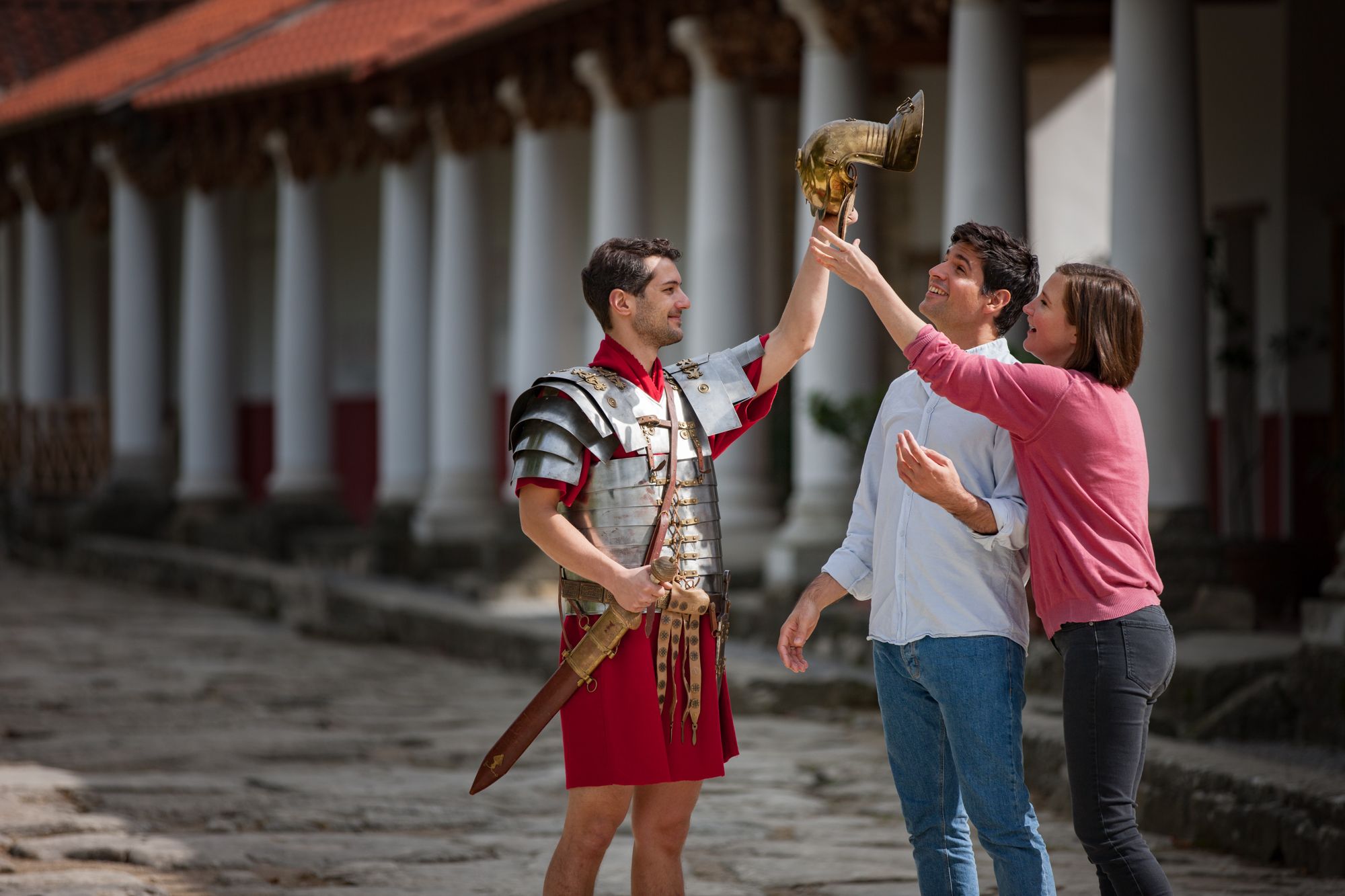 Ein Mann in römischer Rüstung hält einen Helm, während zwei Personen ihn bewundern.