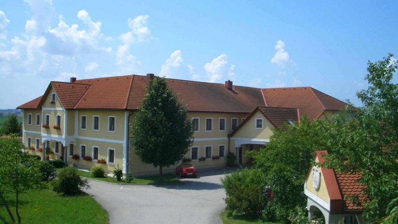 Mayrhof, © zVg Großer Bauernhof mit gelben Wänden und roten Dächern, umgeben von Bäumen und einer Straße.