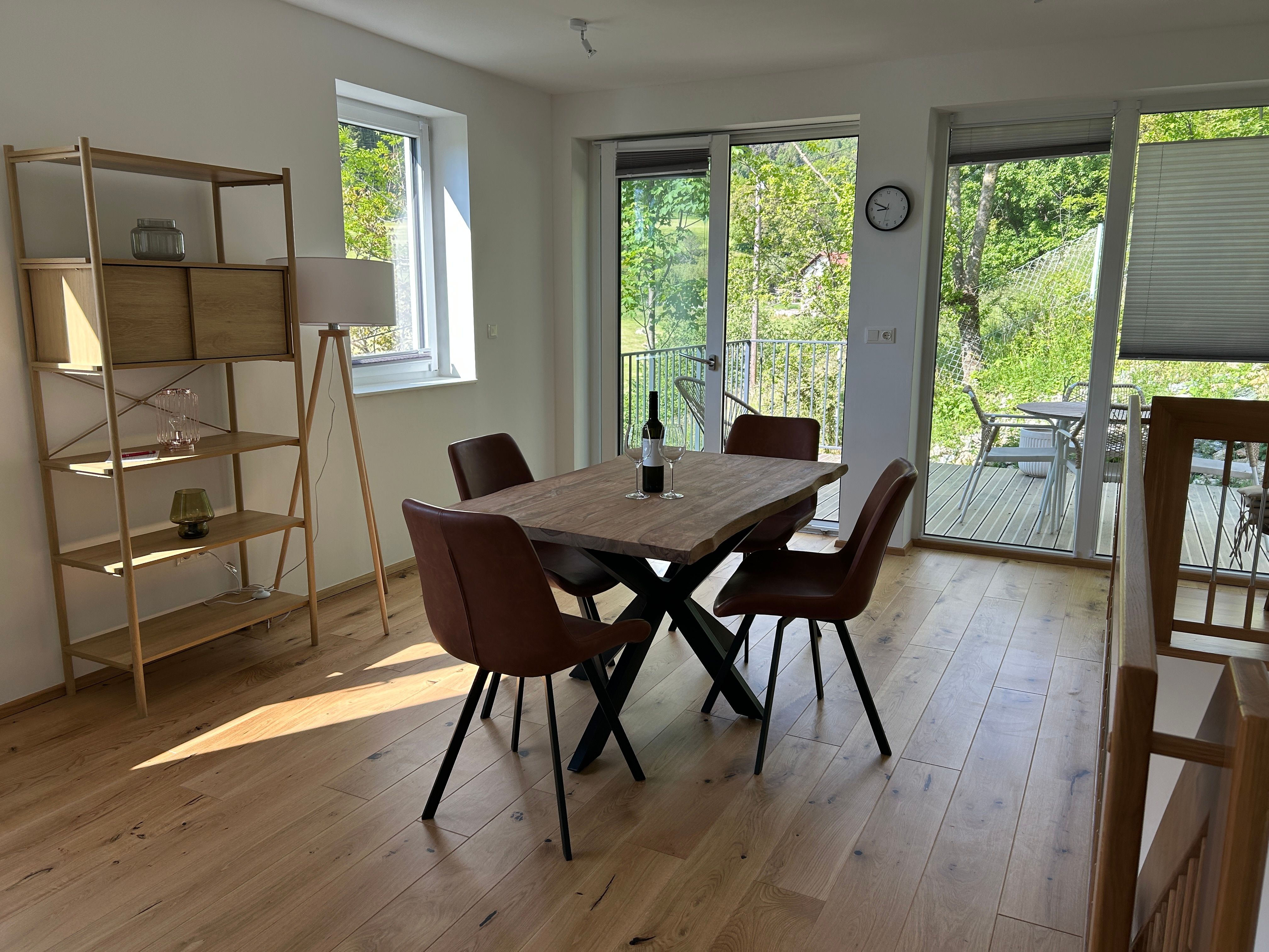 Innenansicht eines modernen Wohnzimmers mit Holzboden, Esstisch und Stühlen, Regal und Blick auf eine Terrasse mit Garten.