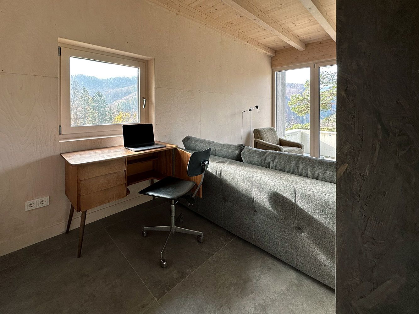 Ein moderner Arbeitsplatz mit einem Laptop auf einem Holztisch, einem schwarzen Stuhl und einem Sofa in einem Raum mit großen Fenstern und Waldblick.