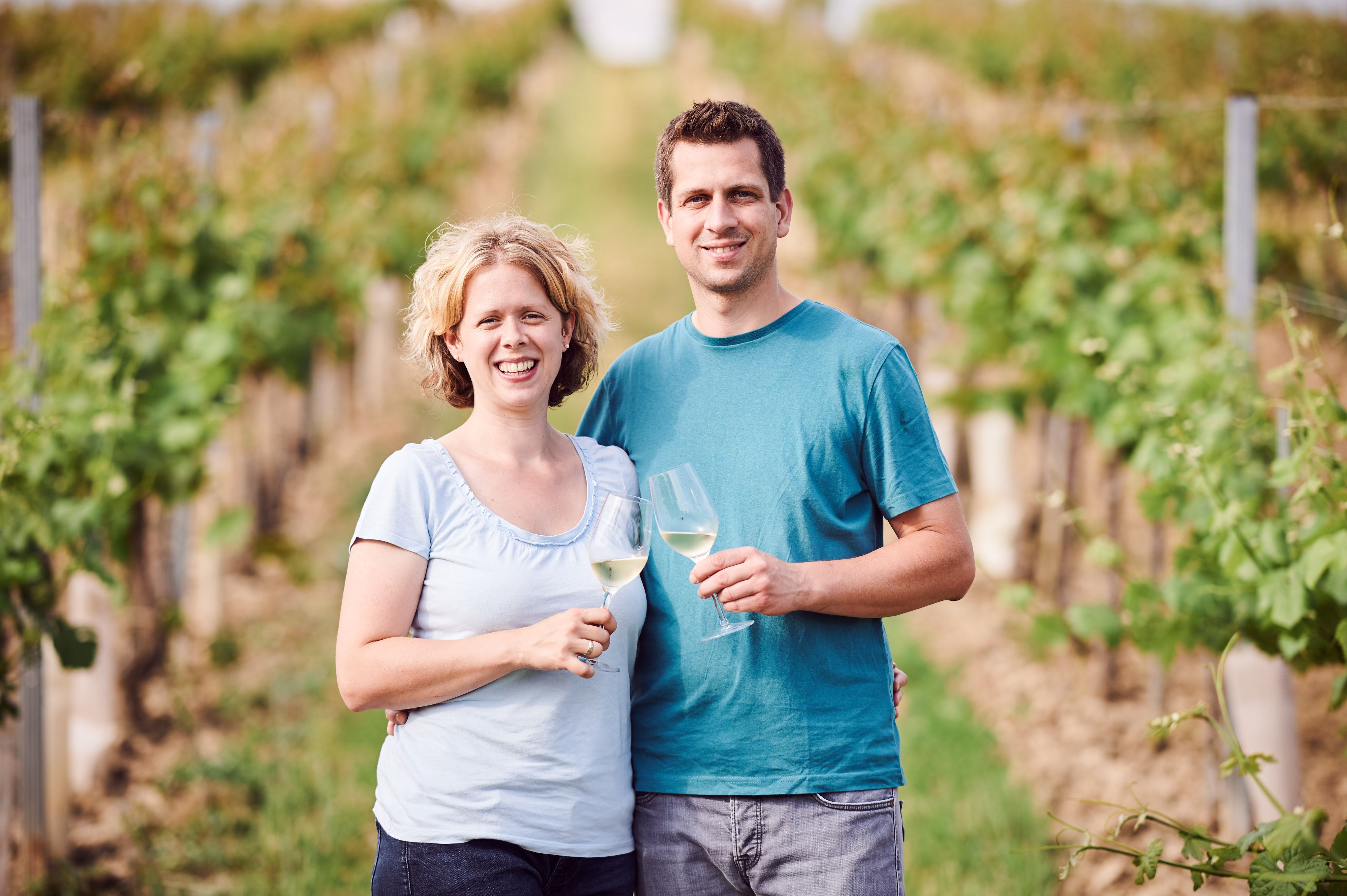 Ein lächelndes Paar steht in einem Weinberg und hält Weingläser in der Hand.