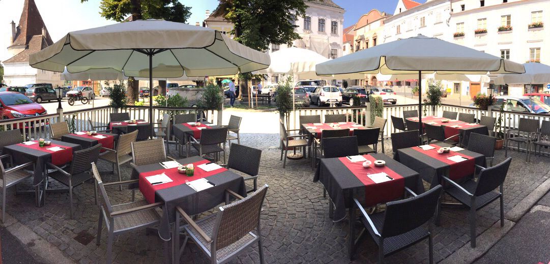 Außenterrasse eines Cafés mit Tischen und Sonnenschirmen auf einem gepflasterten Platz.