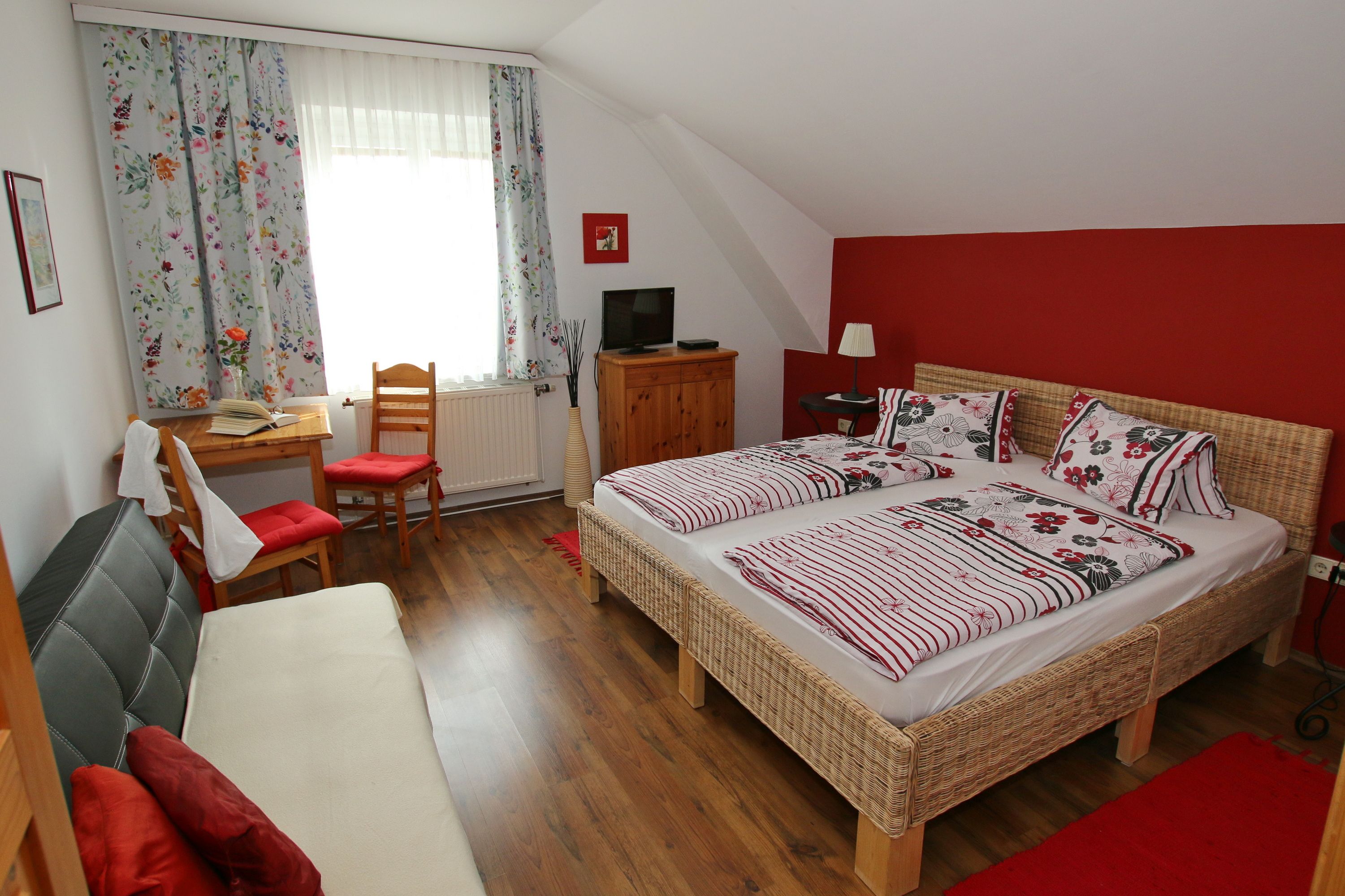 Gemütliches Gästezimmer mit Doppelbett, Sofa, Tisch und Stühlen. Rote Akzentwand und florale Vorhänge.