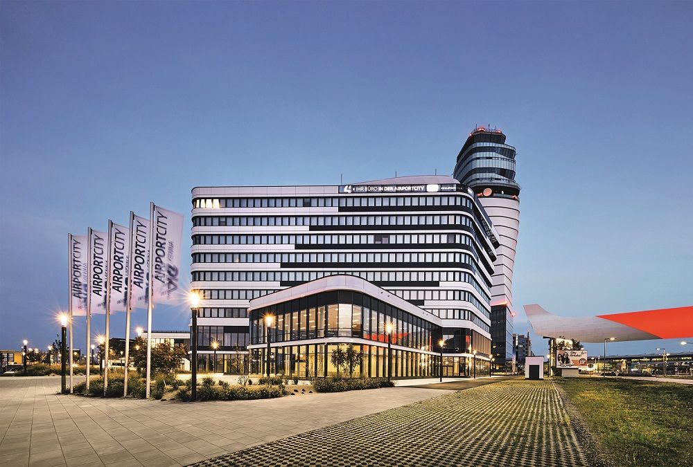Modernes Gebäude am Flughafen Wien bei Dämmerung mit Flaggen und Kontrollturm im Hintergrund.