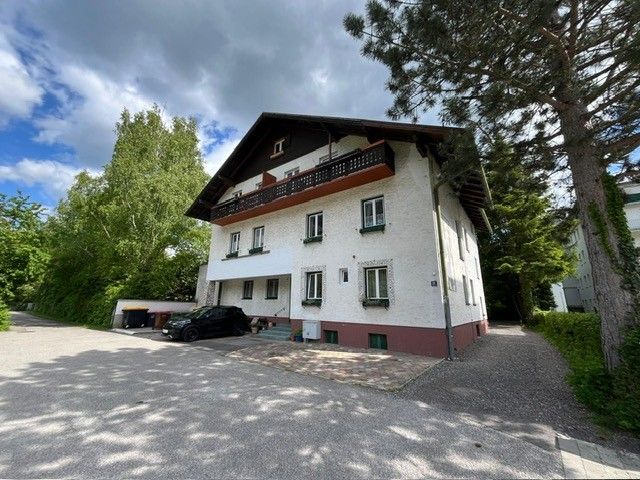 Zweistöckiges Haus mit weißer Fassade und dunklem Dach, umgeben von Bäumen und einem gepflasterten Hof.
