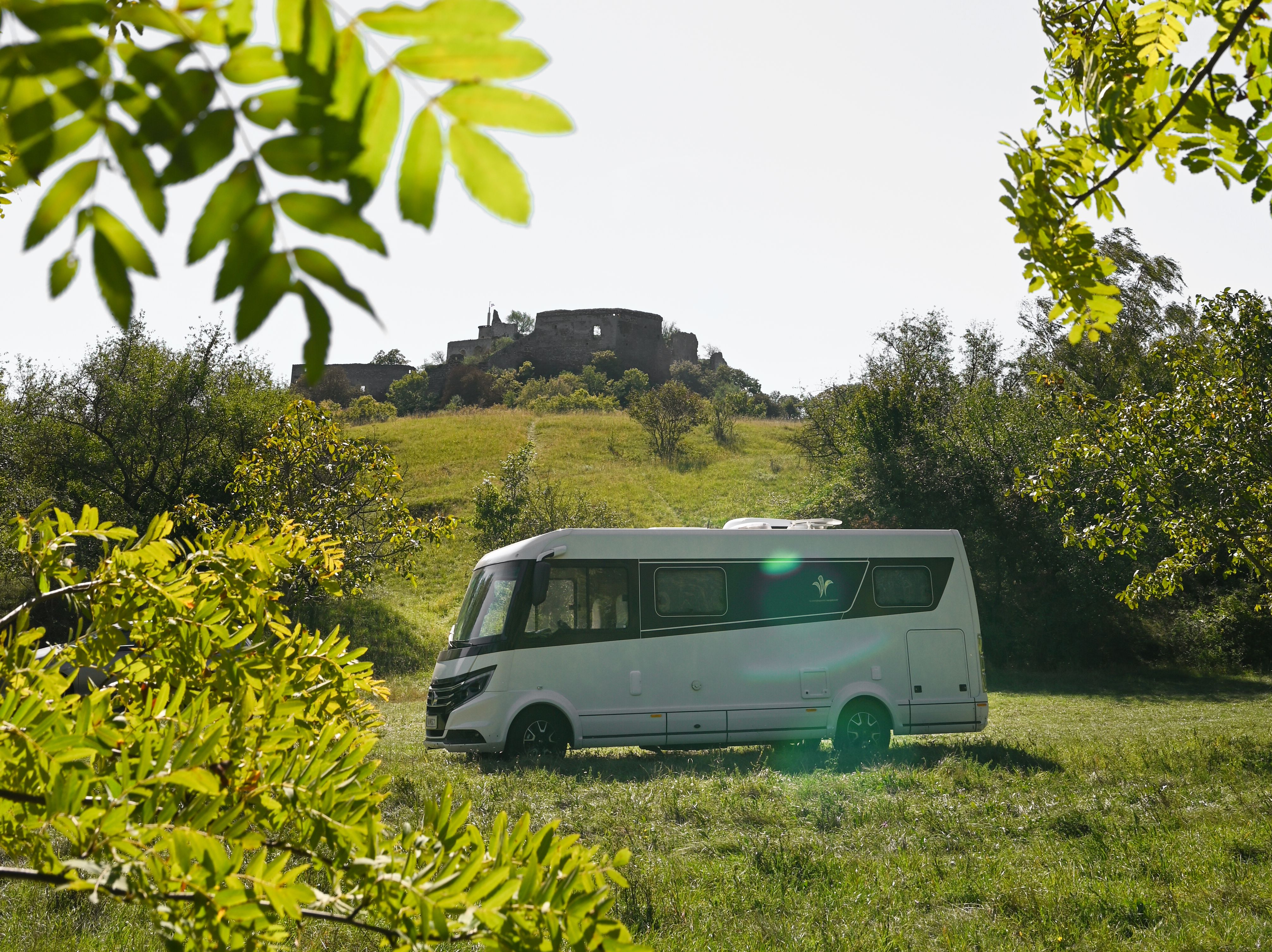 Umgeben von üppigem Grün und unter einem strahlend blauen Himmel, bietet der Campingwagen einen perfekten Rückzugsort für Naturliebhaber. Die sanften Hügel im Hintergrund und die historische Burgruine schaffen eine malerische Kulisse, die zum Entspannen und Erkunden einlädt.