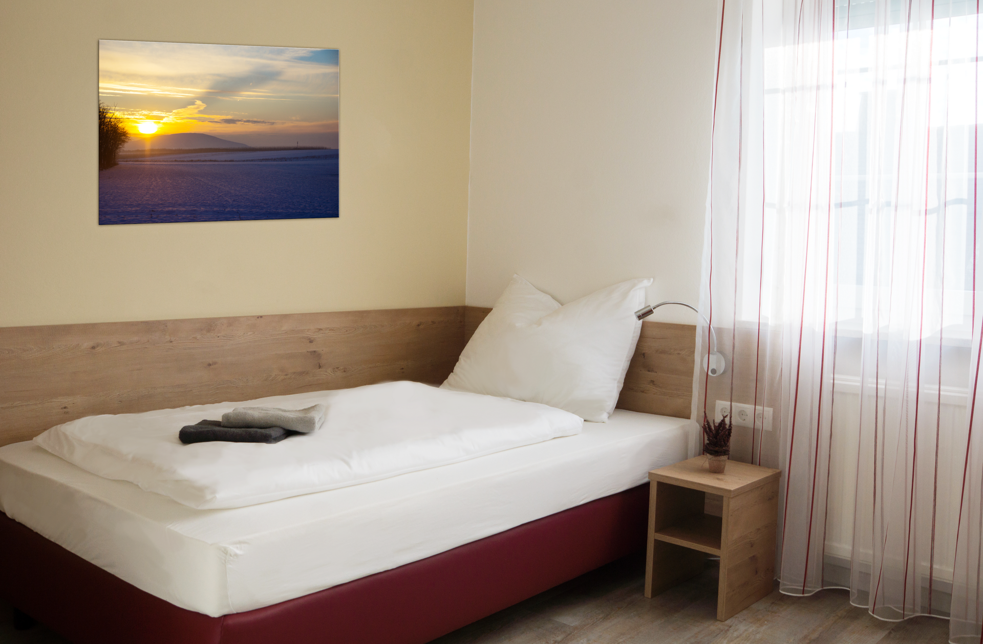 Ein schlichtes Einzelzimmer mit einem Bett, einem Nachttisch und einem Bild an der Wand. Ein Fenster mit Vorhängen lässt Licht herein.
