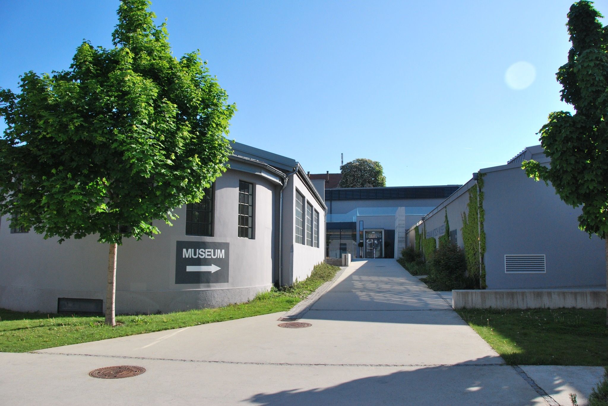 Eingang zum MAMUZ Museum Mistelbach mit Weg zwischen modernen Gebäuden und Bäumen unter blauem Himmel.
