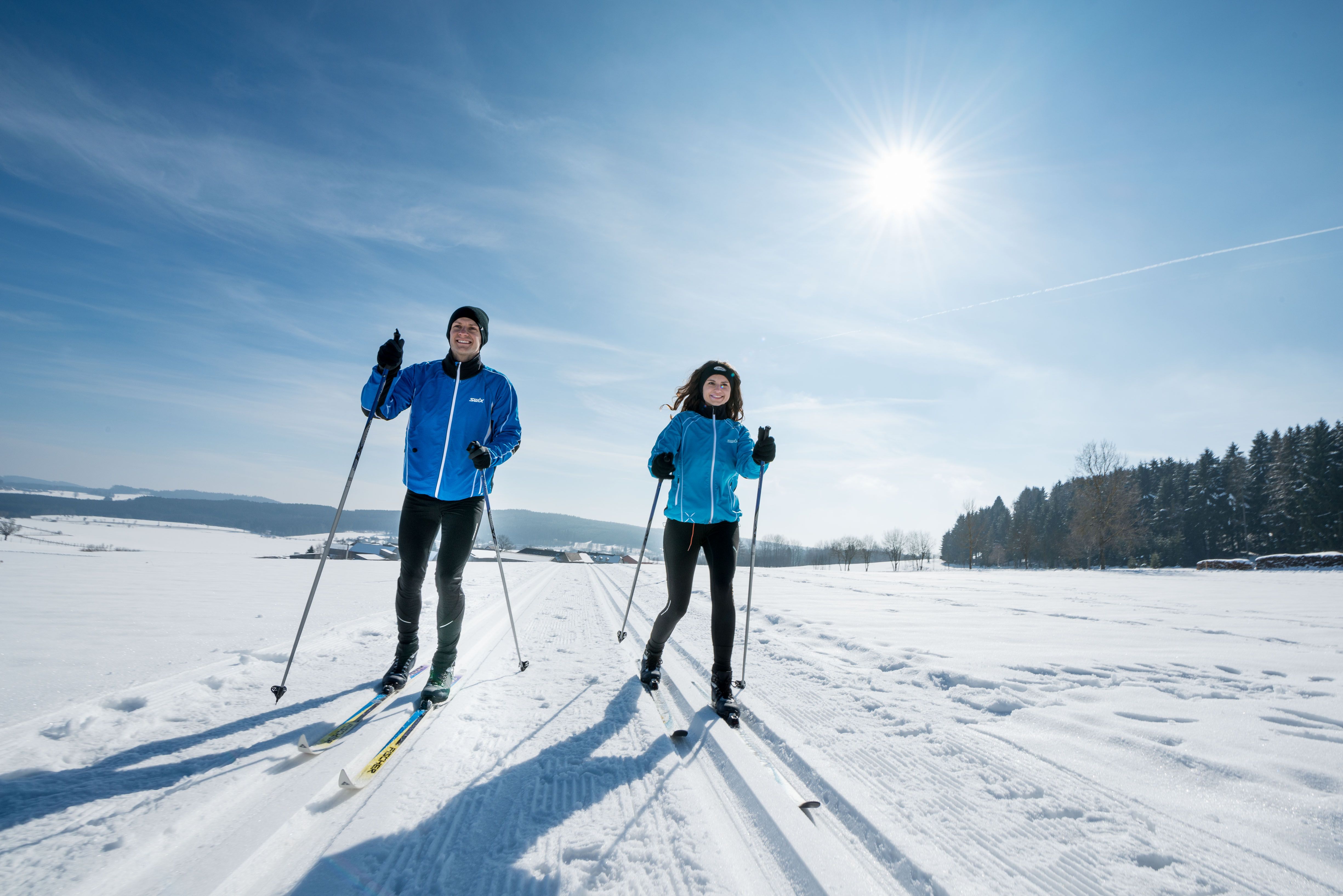 Zwei Personen beim Langlaufen auf einer verschneiten Landschaft unter blauem Himmel und Sonnenschein.