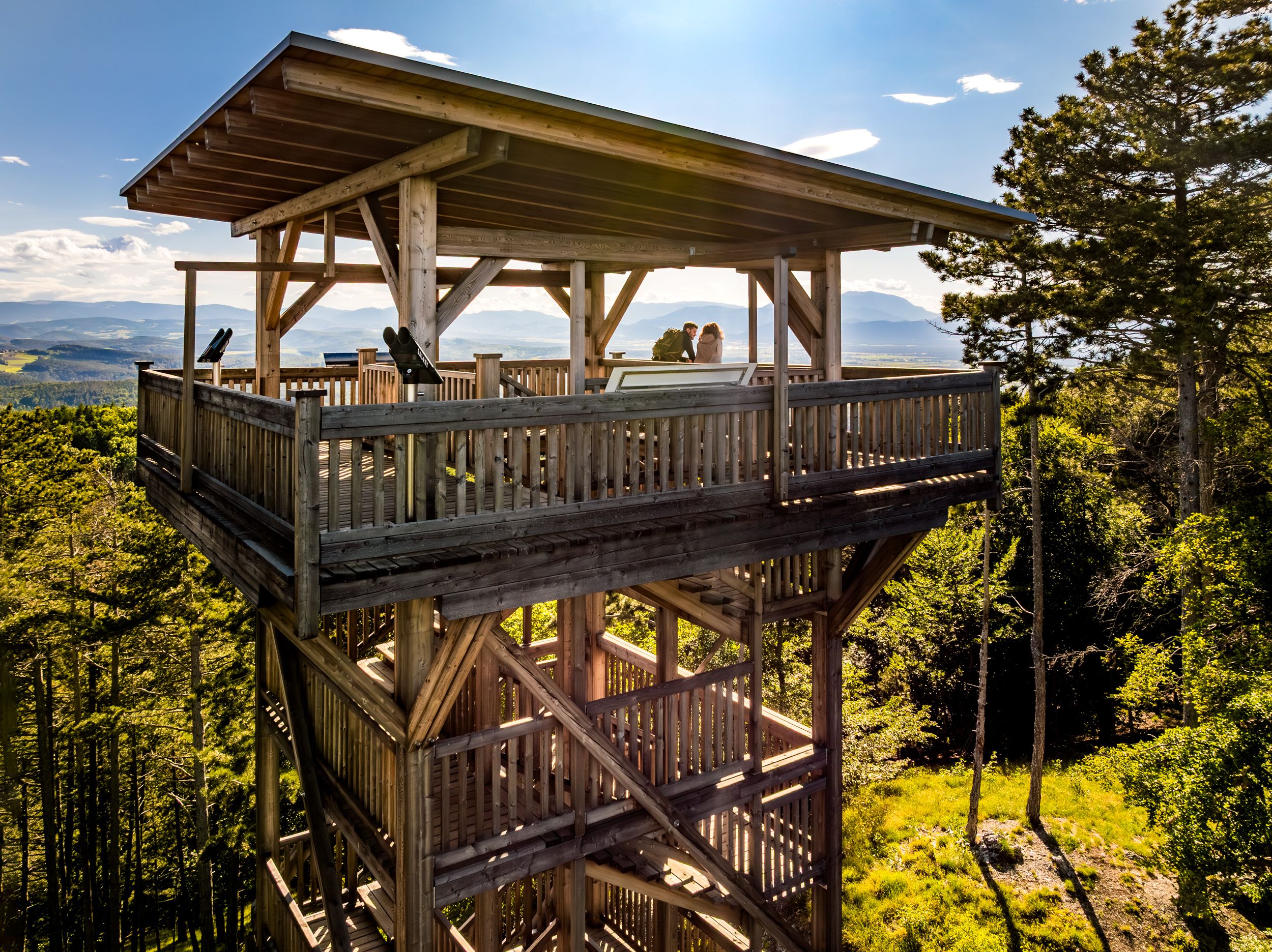 Eine Drohnenaufnahme von der Seite des Aussichtsturmes, auf dem zwei Personen stehen. Im Hintergrund grüner Wald und Berge