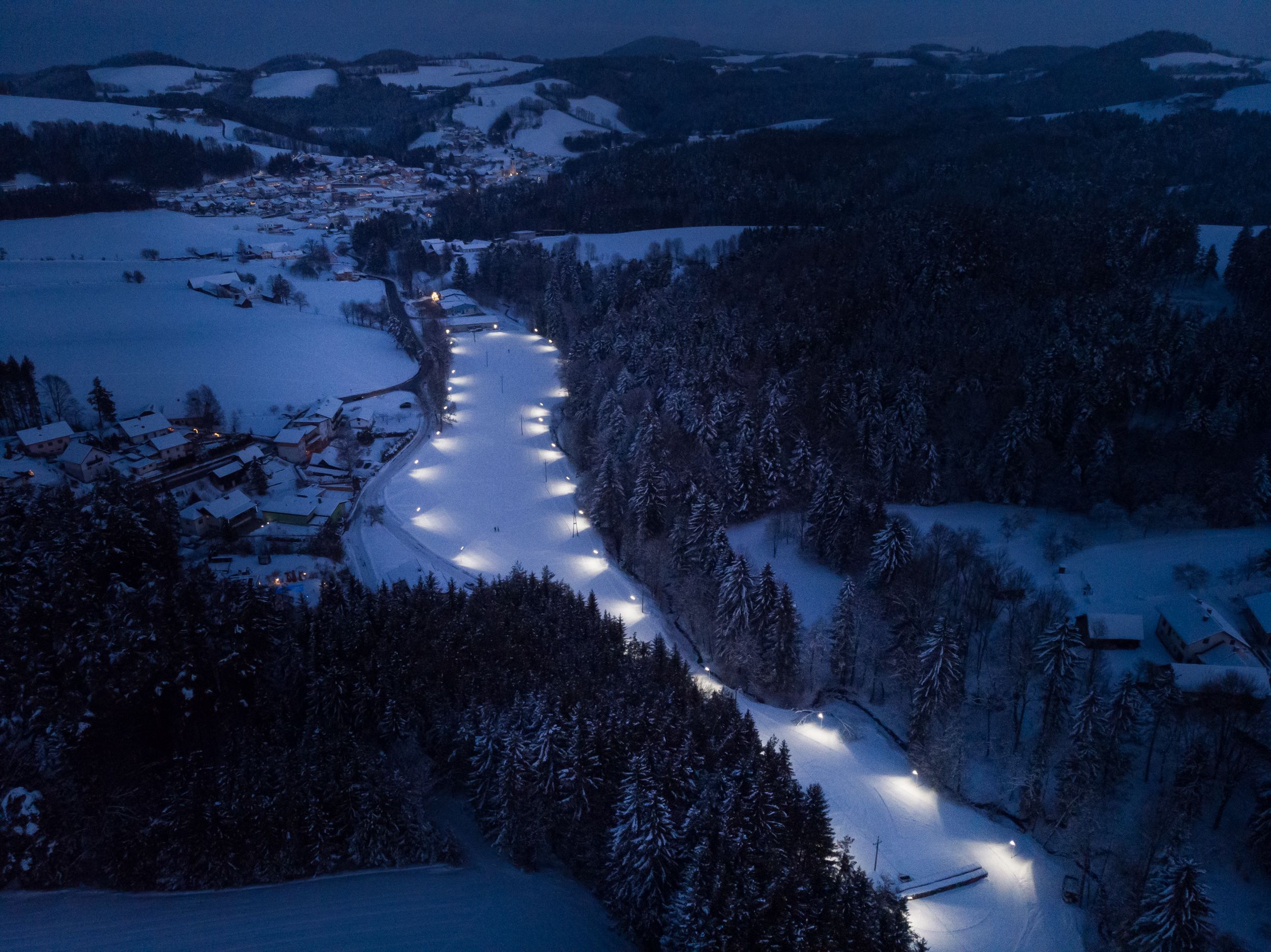 Luftaufnahme einer beleuchteten Langlaufloipe in einer verschneiten Landschaft bei Nacht.