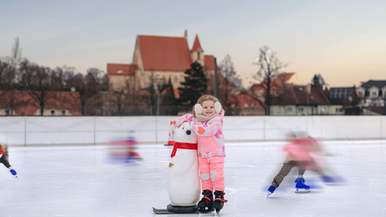 Ein Kind in pinker Kleidung steht auf einer Eisbahn mit einem Pinguin-Hilfsmittel. Im Hintergrund ist eine Kirche zu sehen.