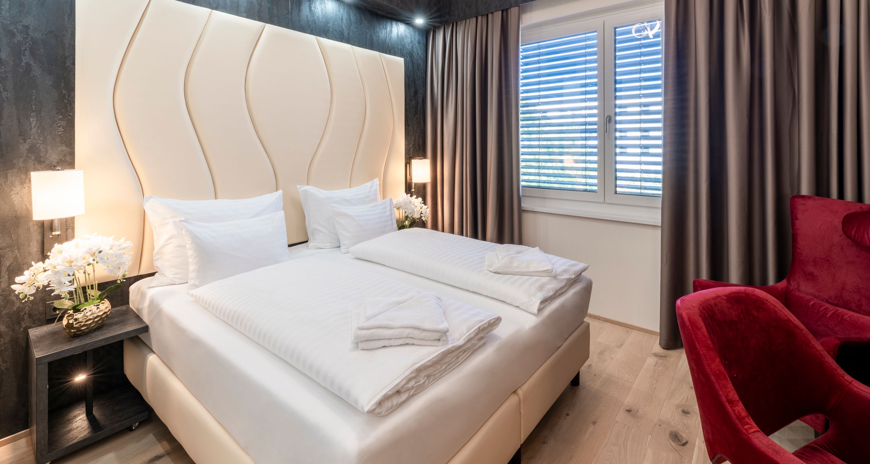 Modernes Hotelzimmer mit Doppelbett, roten Sesseln und großen Fenstern mit Vorhängen.