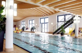 Innenansicht eines Schwimmbads mit Holzdecke und großen Fenstern.