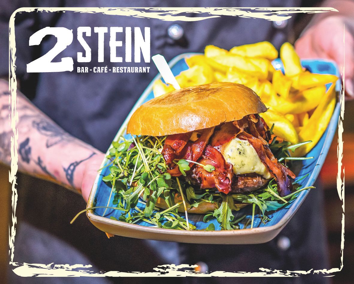Ein Burger mit Speck, Käse und Rucola auf einem Teller mit Pommes, präsentiert von einer tätowierten Hand. Logo von 2Stein Bar-Café-Restaurant oben links.