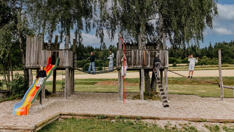 Kinder spielen auf einem h&ouml;lzernen Spielplatz mit Rutsche und Seilbr&uuml;cke im Freien.