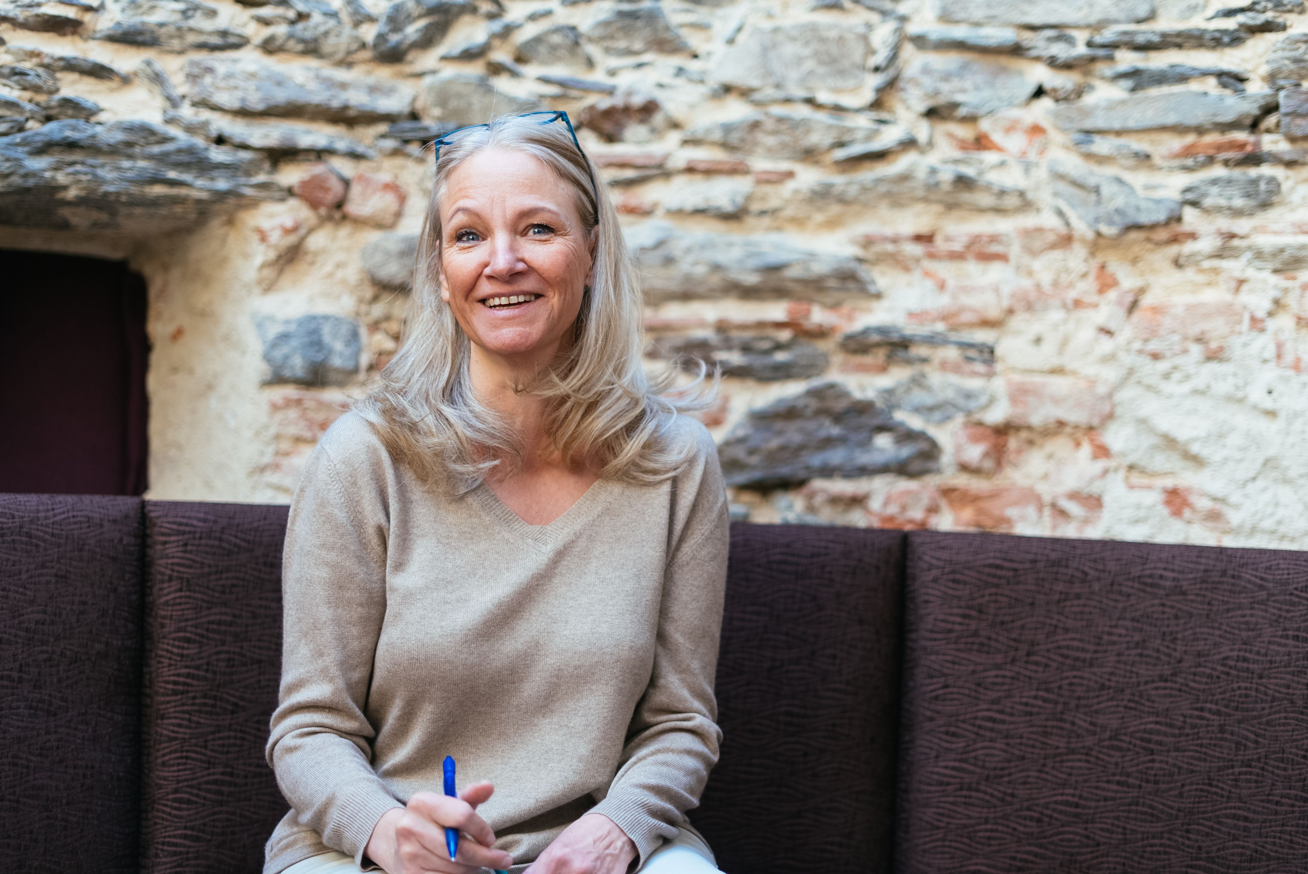 Eine grauhaarige Frau sitzt lächelnd auf einer gepolsterten Bank vor einer rustikalen Natursteinmauer. Sie trägt einen hellen Pullover, hat eine Brille am Kopf und hält einen blauen Stift in der Hand.
