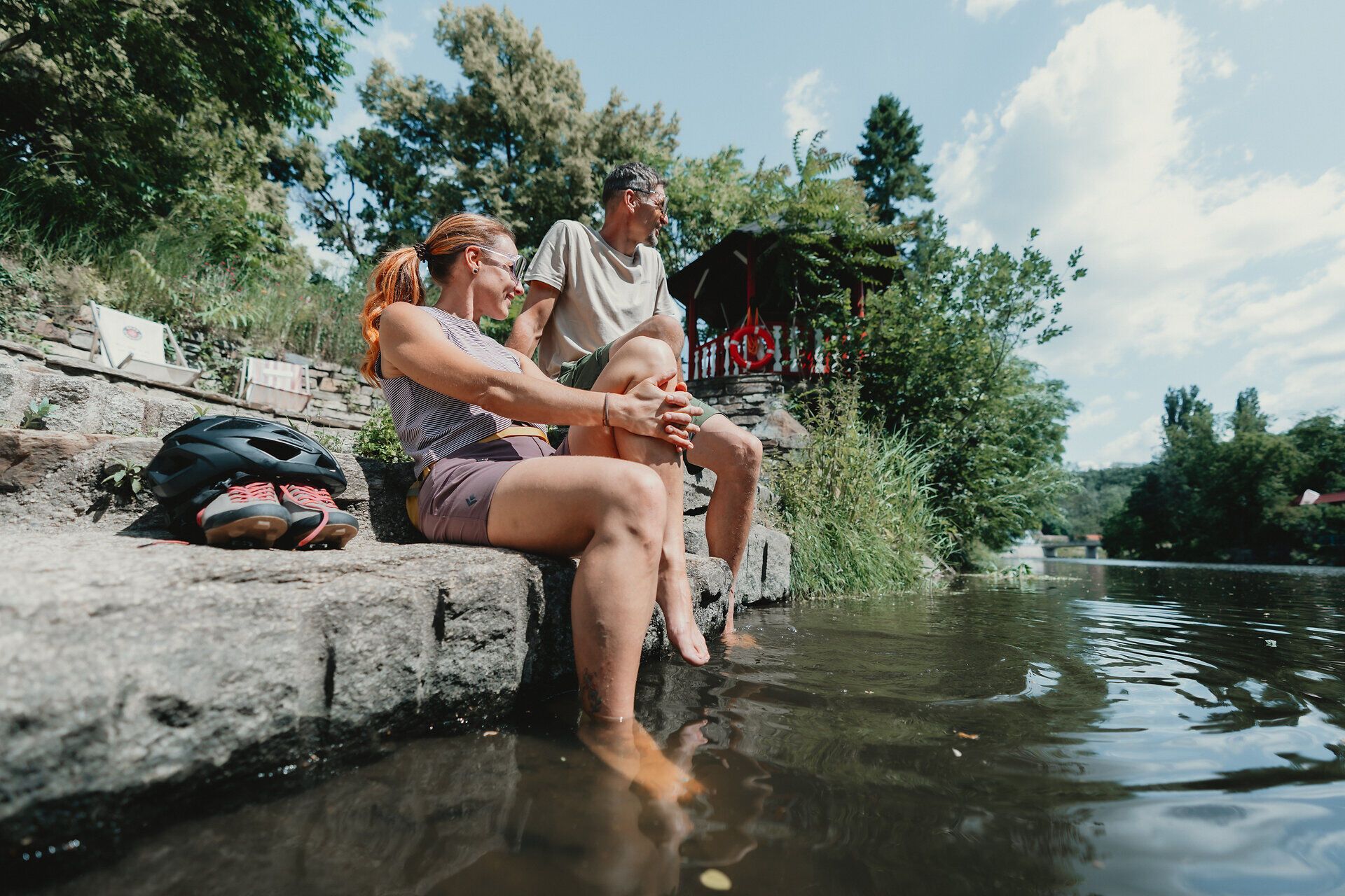 Zwei Personen sitzen auf einer Steintreppe am Flussufer und kühlen ihre Füße im Wasser.