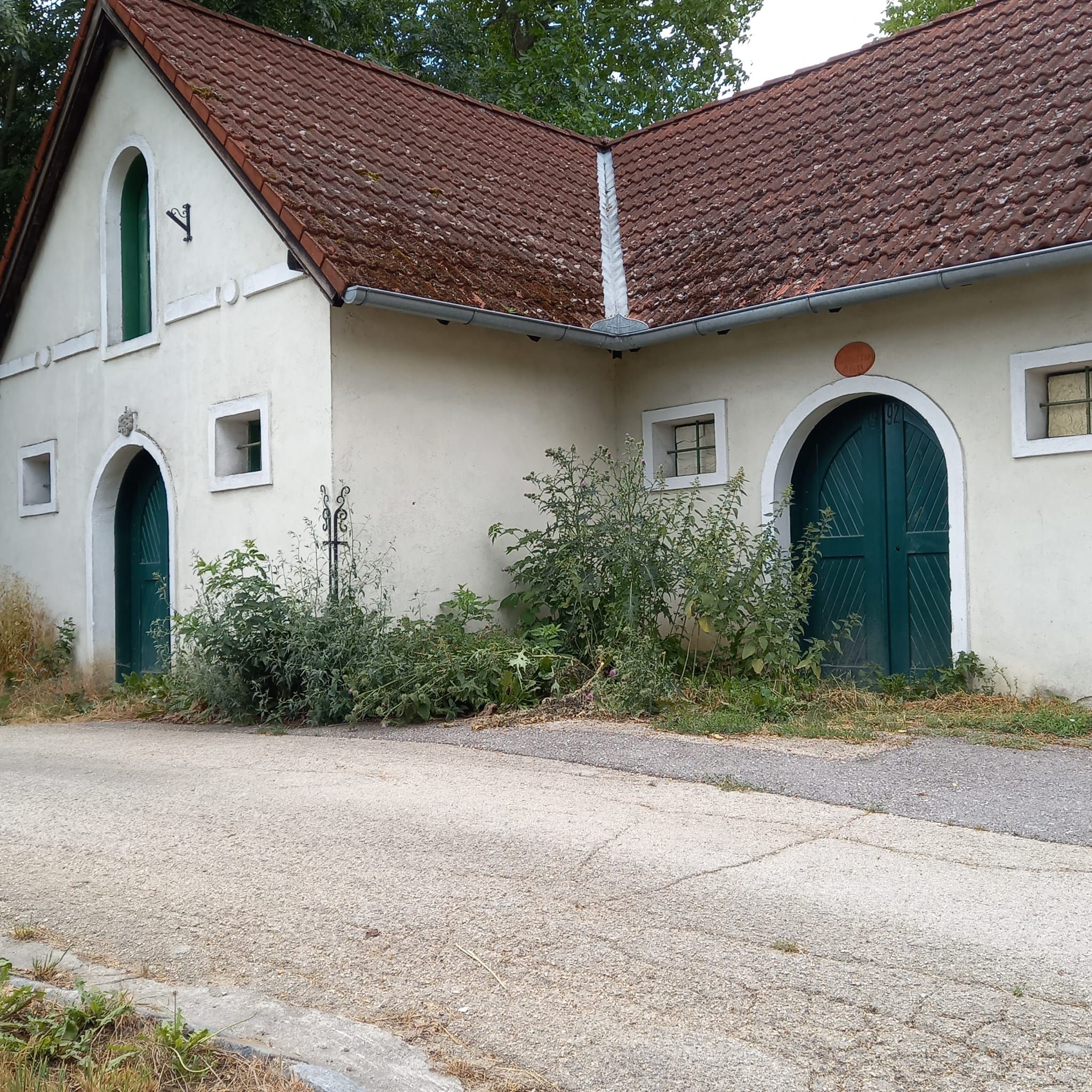 Altes Gebäude mit roten Ziegeldach und grünen Türen, umgeben von Pflanzen.