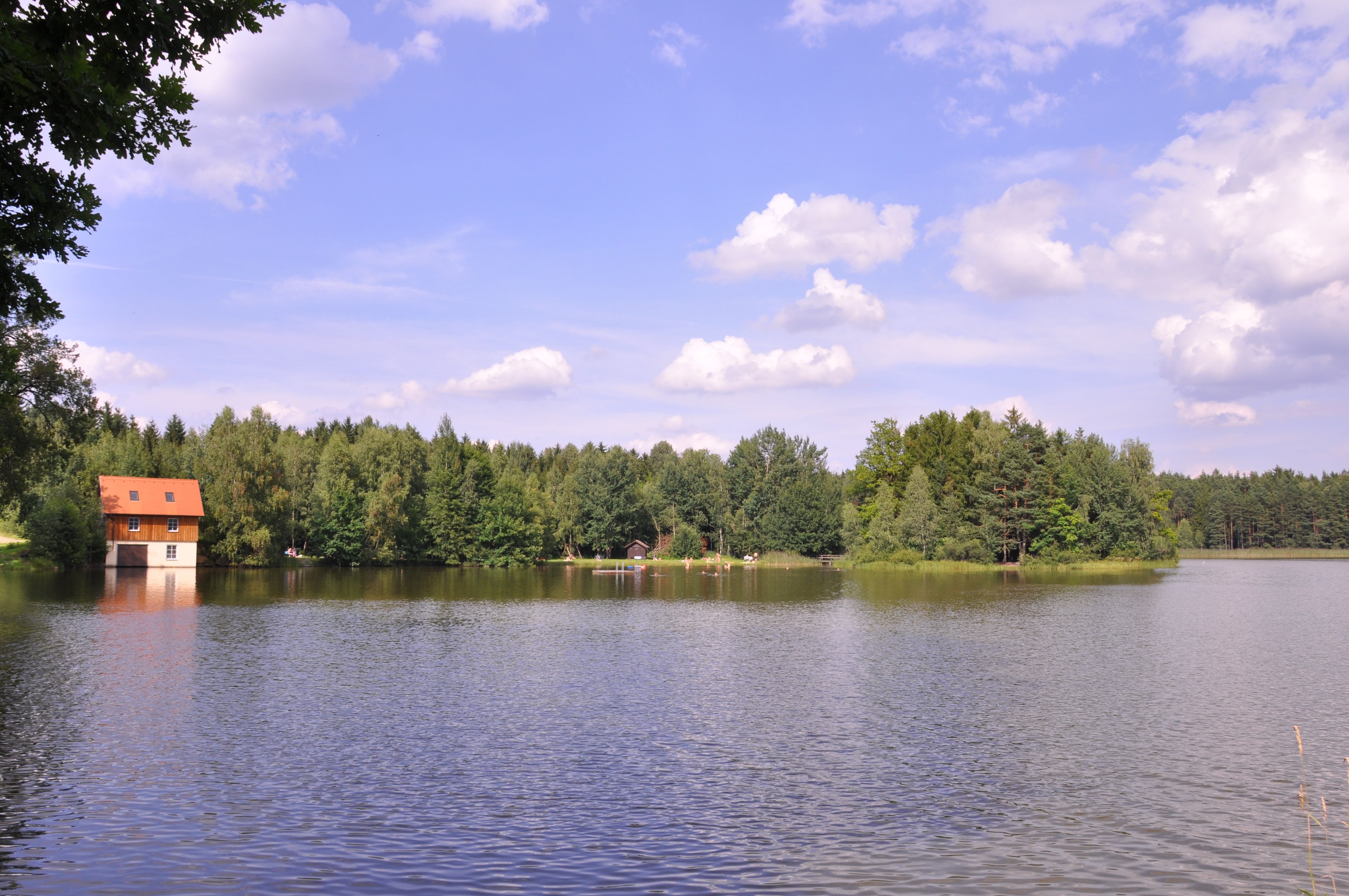 Ein See mit einem Haus am Ufer, umgeben von Wald und blauem Himmel mit Wolken.