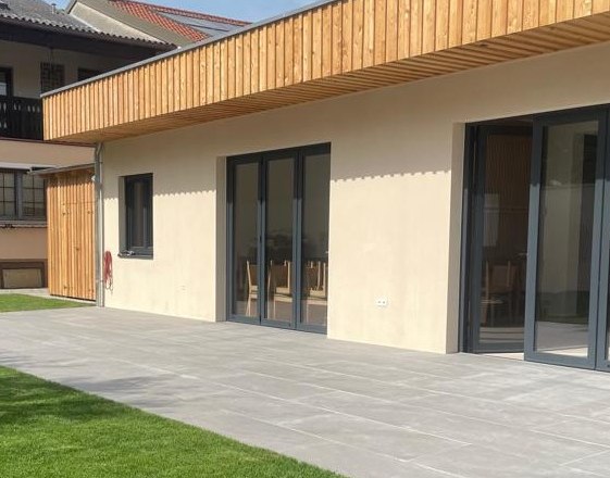 Moderne Gebäudeaußenansicht mit Holzverkleidung und großen Fenstern, umgeben von einer Terrasse und Rasenfläche.