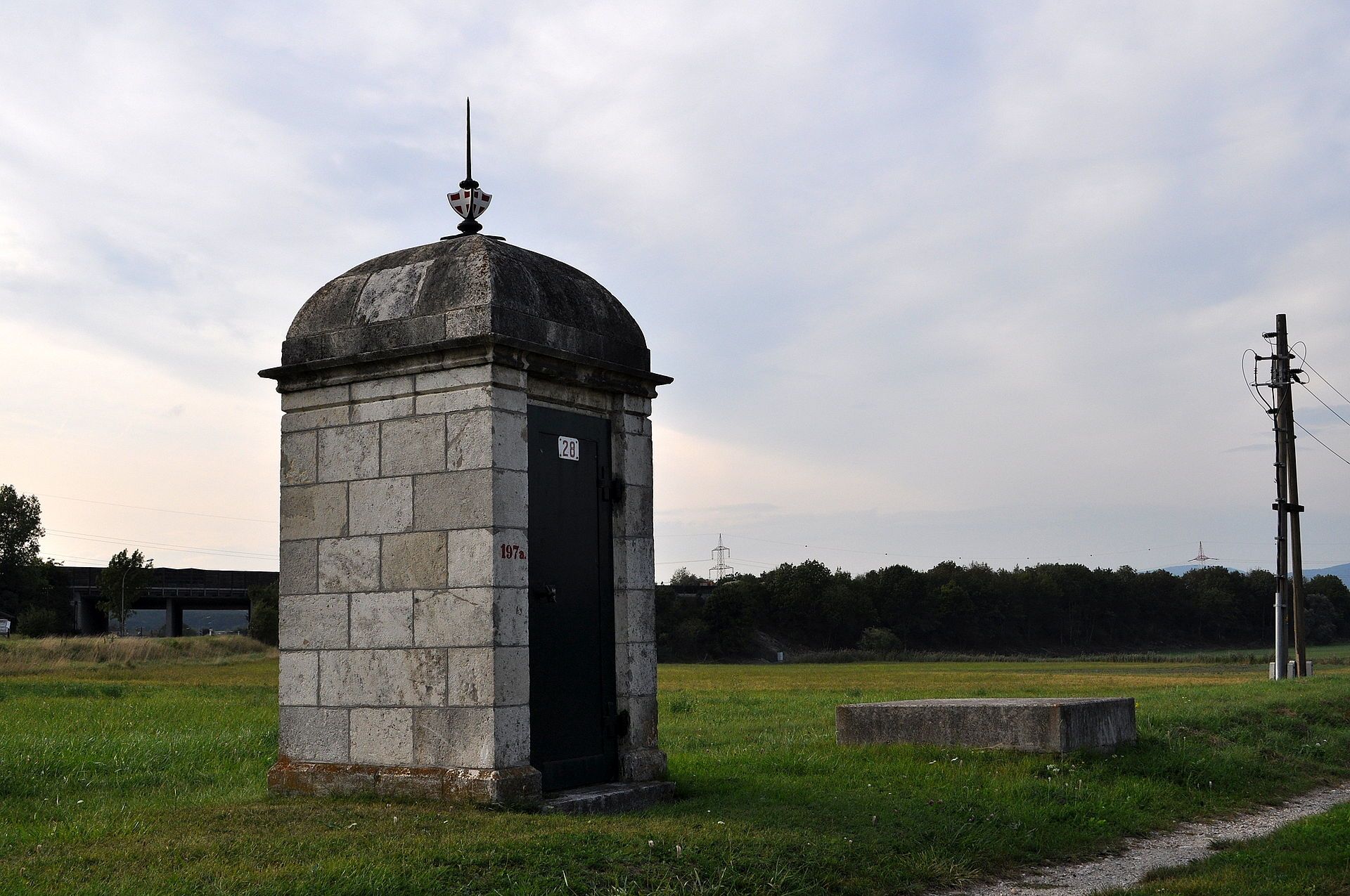 Steinbauwerk auf einer Wiese mit Strommast im Hintergrund.