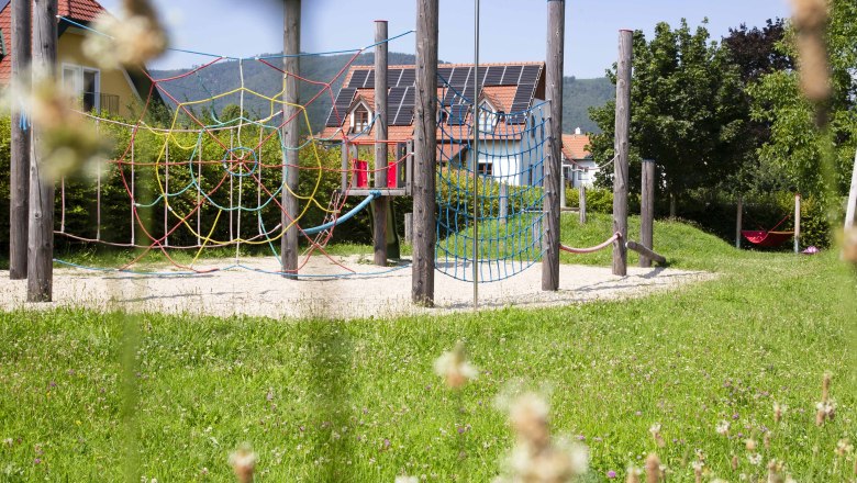 Ein Abenteuerspielplatz mit Kletternetzen und Holzpfosten, umgeben von gr&uuml;ner Wiese und H&auml;usern im Hintergrund.