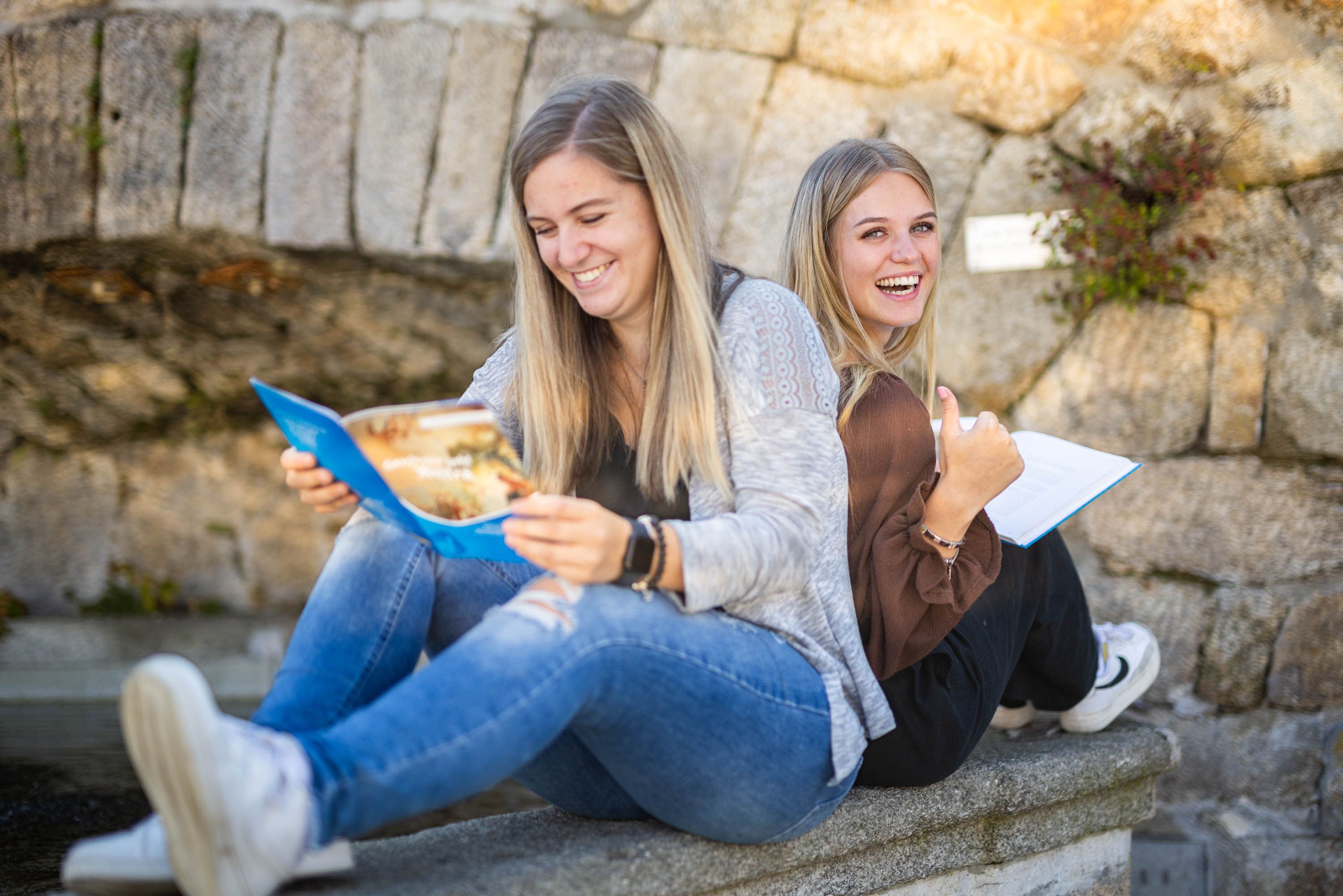Zwei lachende Frauen sitzen Rücken an Rücken auf einer Steinmauer und lesen Broschüren.