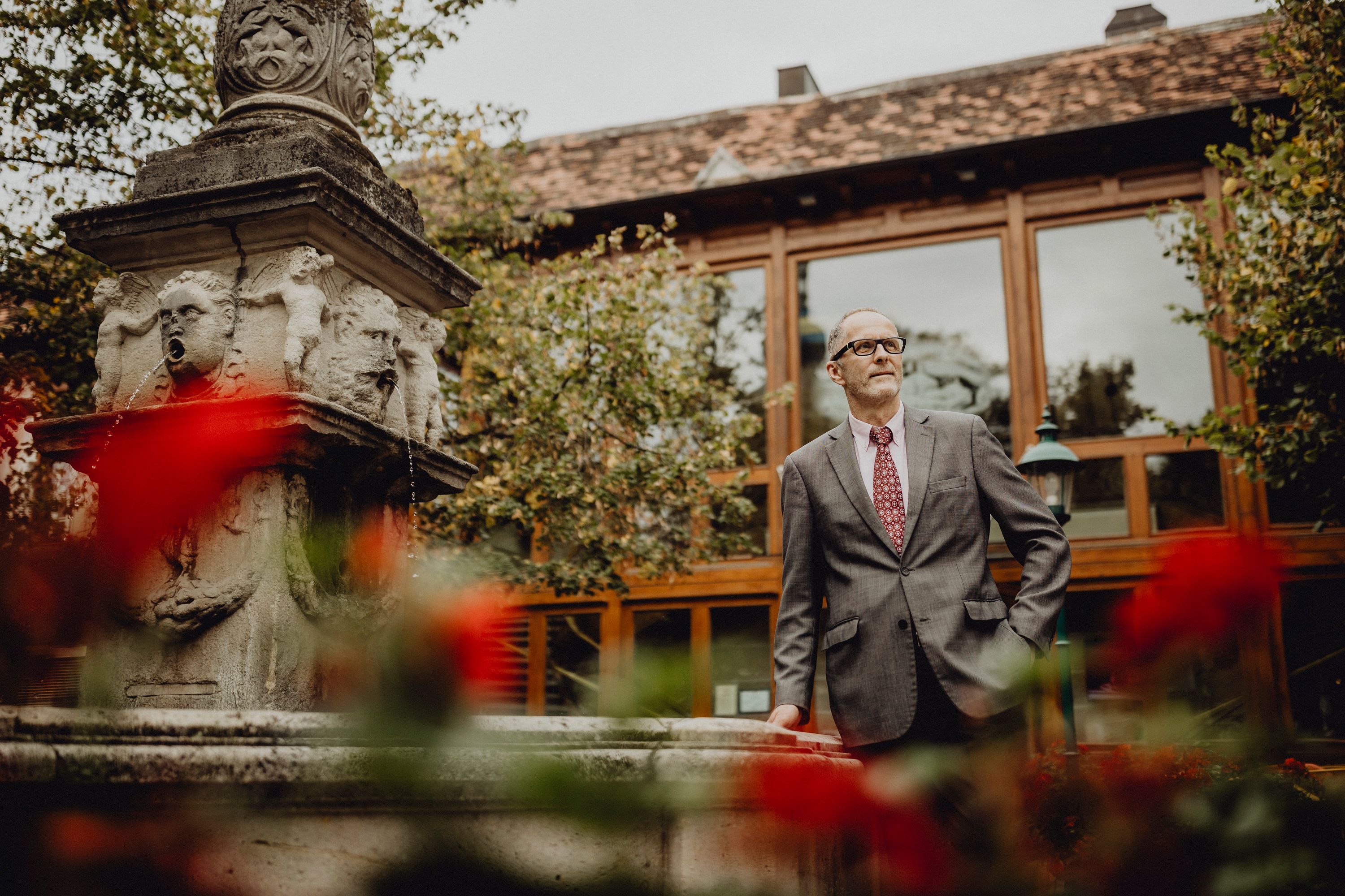 Ein Mann im Anzug steht neben einem steinernen Brunnen mit Wasserspeiern, umgeben von roten Blumen und einem Gebäude im Hintergrund.