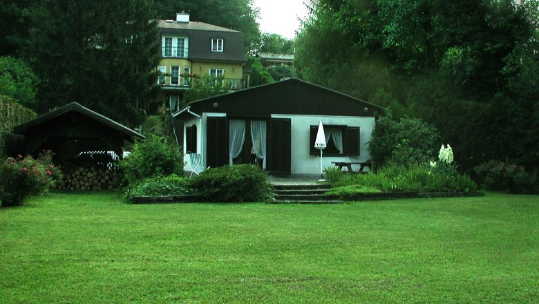 Haus, © Haus Ryba Ein kleines Haus mit Garten und Terrasse, umgeben von Bäumen und einem größeren Gebäude im Hintergrund.