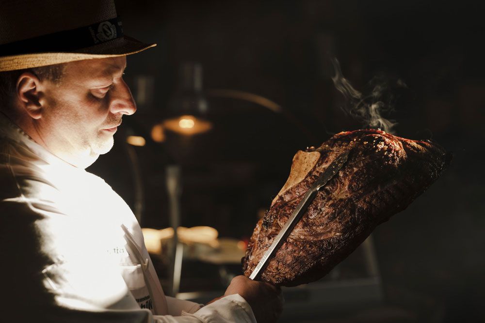 Ein Mann in einem weißen Hemd hält ein großes, gegrilltes Stück Fleisch mit einem Messer.