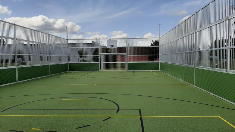 Ein leerer, eingez&auml;unter Multisportplatz mit gr&uuml;nem Boden und Markierungen f&uuml;r verschiedene Sportarten unter einem blauen Himmel mit Wolken.