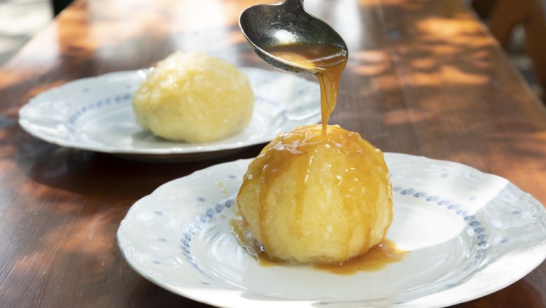 Ein Knödel auf einem Teller wird mit Soße übergossen, ein weiterer Knödel im Hintergrund.