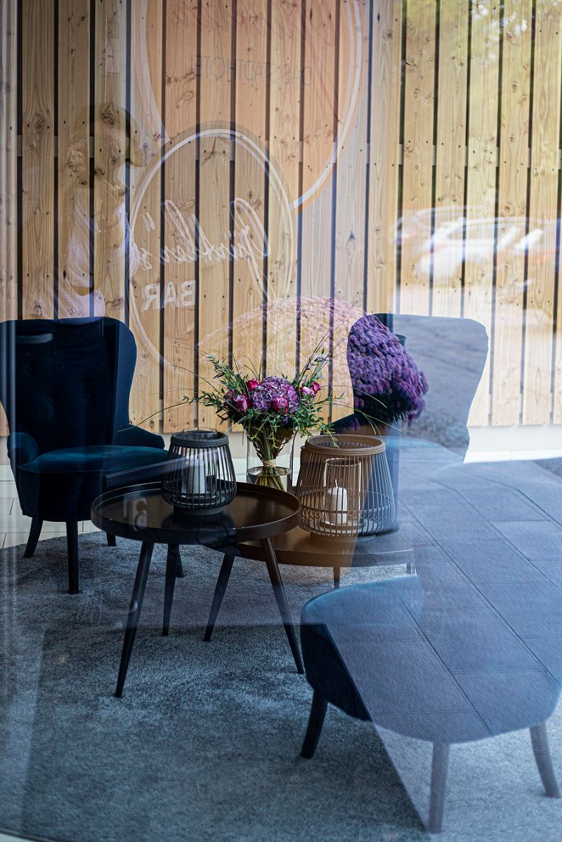 Ein gemütlicher Loungebereich mit blauen Sesseln, einem Tisch mit Kerzen und einem Blumenstrauß, reflektiert in einer Glaswand.