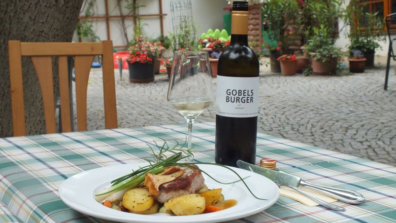 Ein Teller mit Fleisch und Kartoffeln auf einem Tisch im Freien, daneben eine Flasche Wein und ein Glas.
