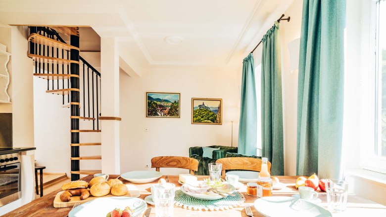 Essbereich mit gedecktem Tisch, Wendeltreppe und gr&uuml;nen Vorh&auml;ngen.