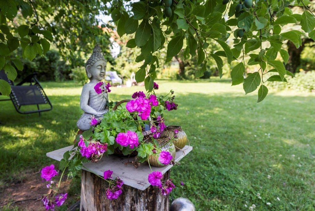 Garten mit Buddha-Statue und pinken Blumen auf einem Holzstumpf.