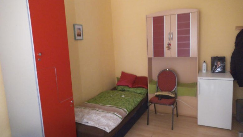 Ein kleines Einzelzimmer mit Bett, rotem Schrank, Stuhl und kleinem K&uuml;hlschrank.