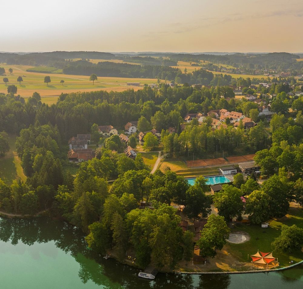 Luftaufnahme eines Sees mit umliegendem Wald und Gebäuden.