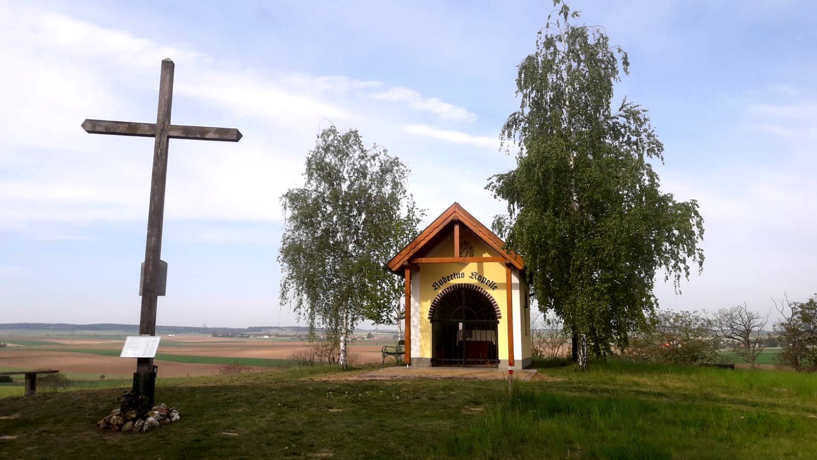 Ein Holzkreuz und eine kleine Kapelle auf einem Hügel mit Bäumen und weitem Blick über die Landschaft.