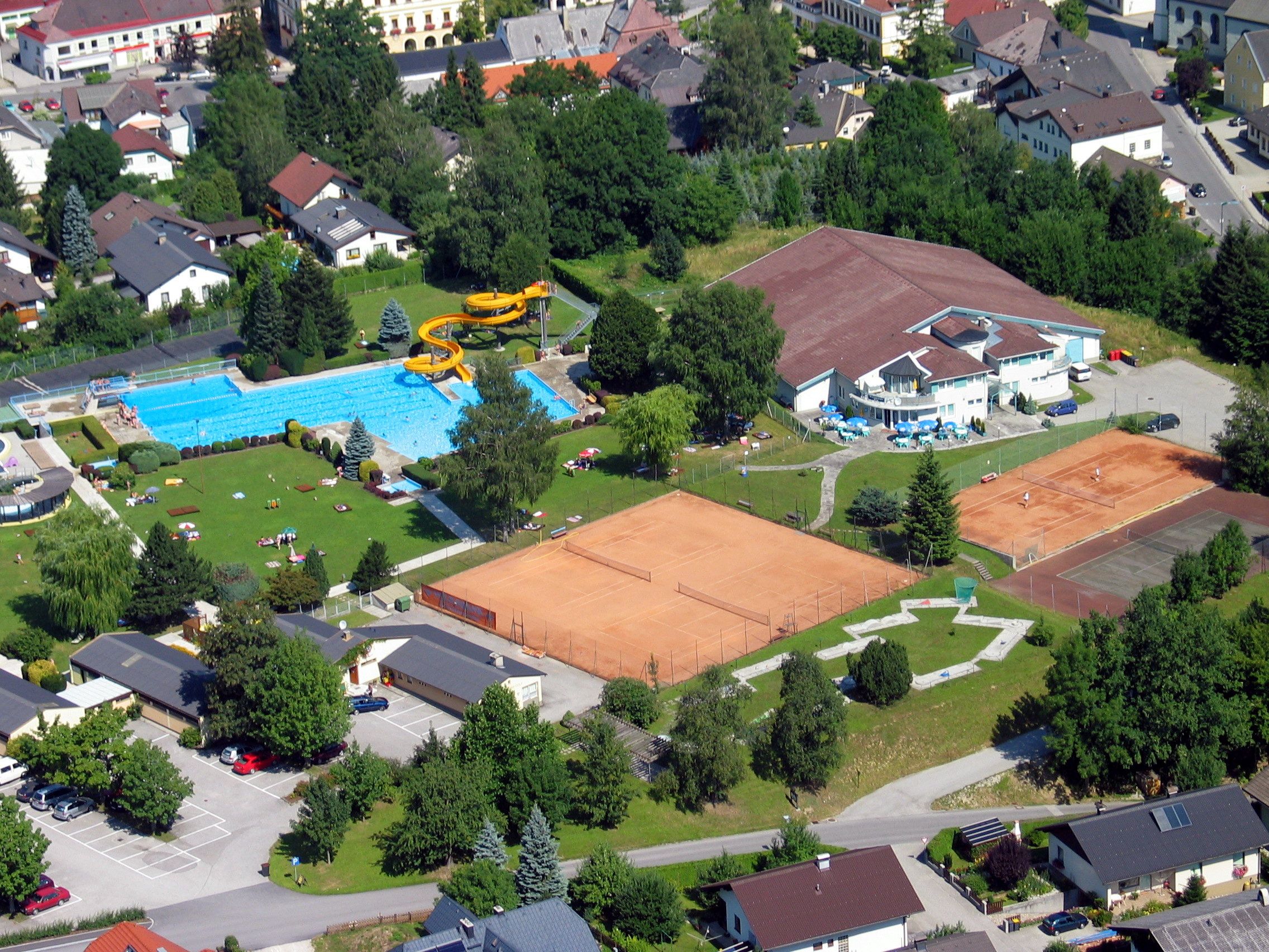 Luftaufnahme eines Freibads mit Schwimmbecken, Rutsche und Tennisplätzen in einer Wohngegend.