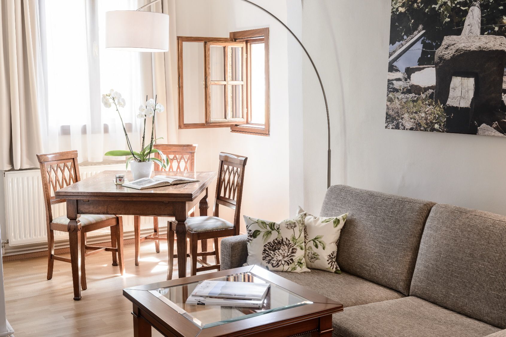 Gemütliches Wohnzimmer mit grauem Sofa, Holztisch und Stühlen, hellem Fenster und Pflanzen.