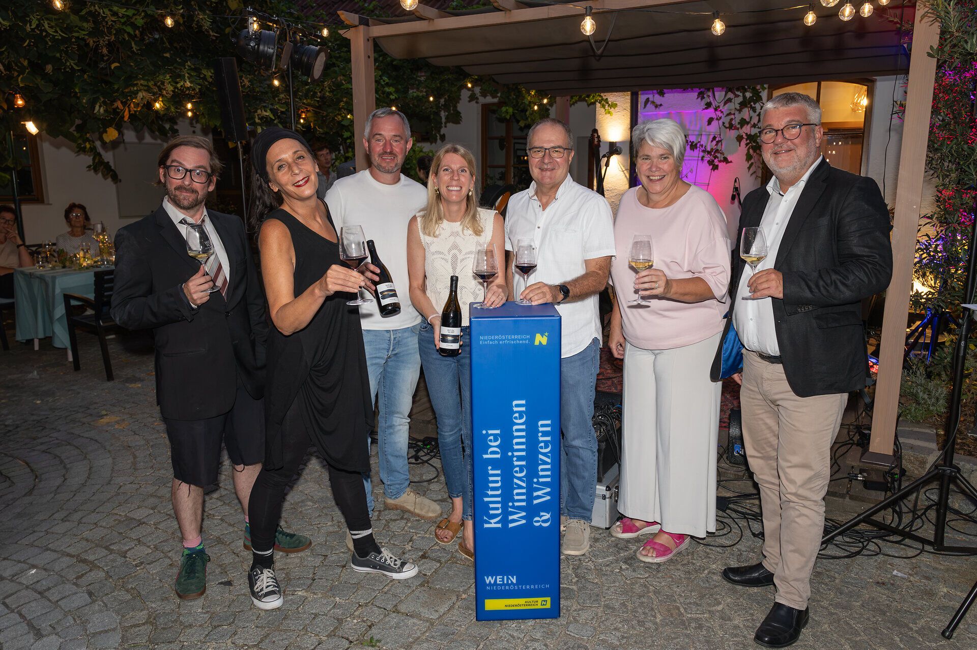Sieben Personen stehen bei einer Abendveranstaltung im Freien mit Weingläsern und Weinflaschen rund um eine Stele mit der Aufschrift „Kultur bei Winzerinnen & Winzern“