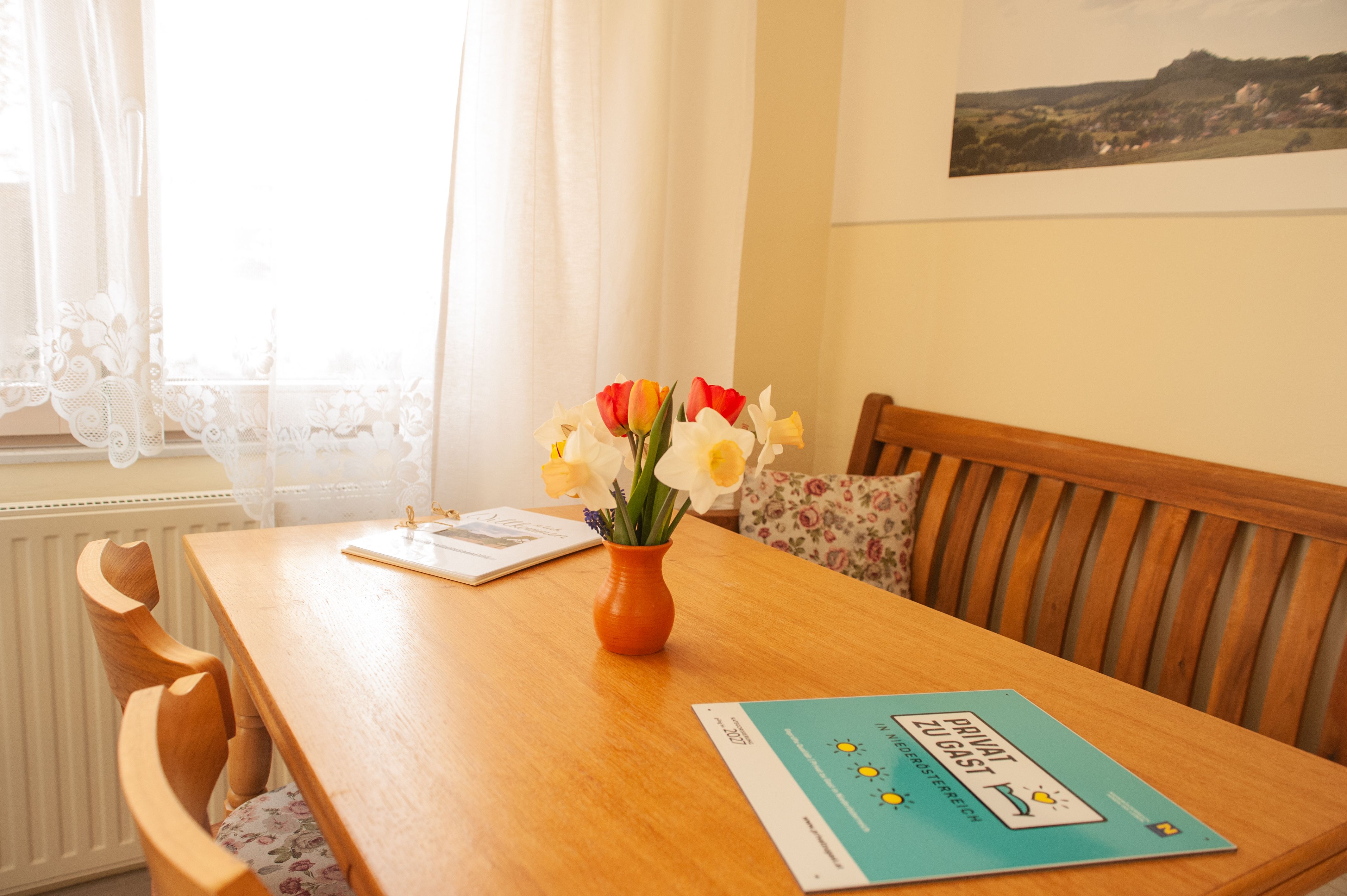 Ein gemütlicher Essbereich mit Holztisch, Blumenstrauß in Vase und Broschüren. Helle Vorhänge und ein Bild an der Wand.