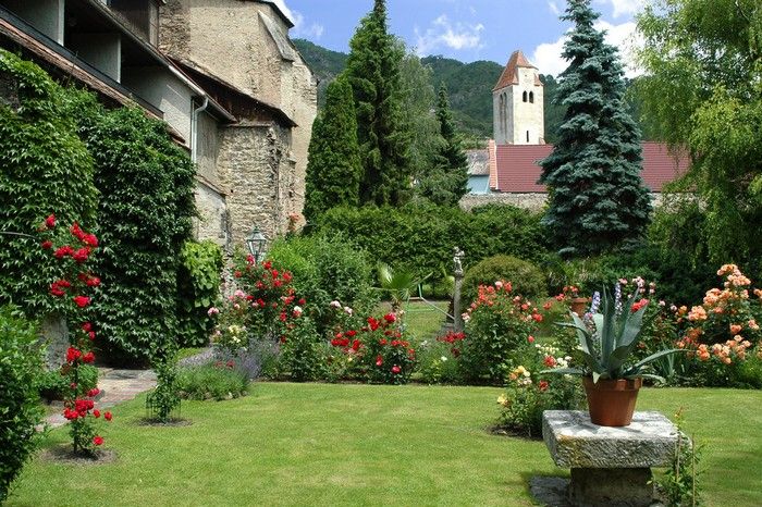 Ein gepflegter Klostergarten mit blühenden Rosen, einem Rasen und einer alten Steinmauer im Hintergrund.