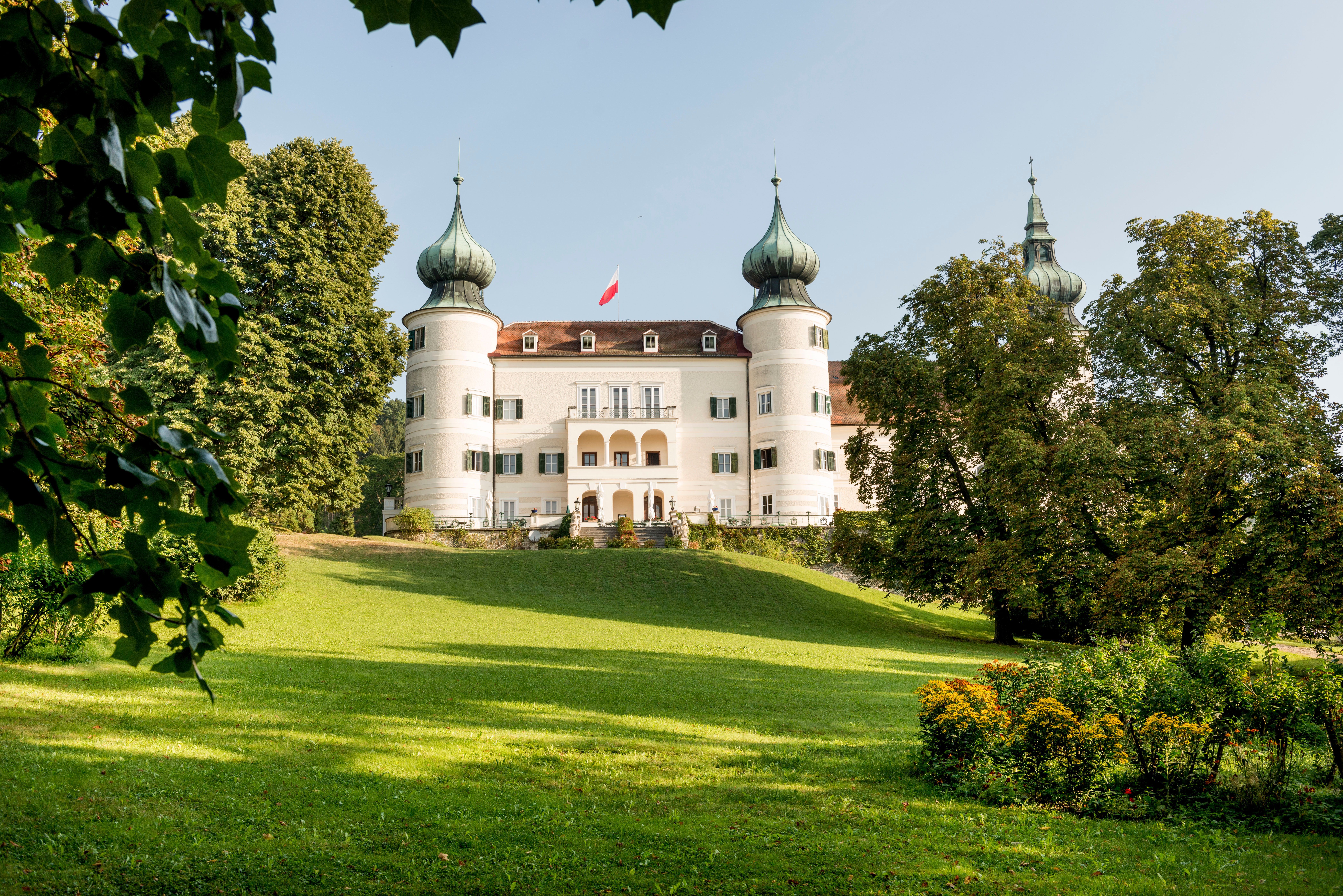 Ein weißes Schloss mit mehreren Zwiebeltürmen, umgeben von grüner Landschaft und Bäumen, unter einem blauen Himmel.
